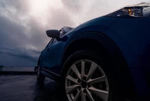 New luxury blue SUV car parked on concrete road beside the beach on rainy day with stormy sky. Front view of blue SUV car with sport design. Raindrops on car. Road trip travel. Driving in bad weather. photo