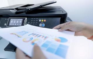 Office worker prints paper on multifunction laser printer. Copy, print, scan, and fax machine in office. Document and paper work. Print technology. Blurred hand hold printed paper. Scanner equipment. photo