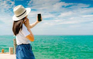 Young Asian woman wear hat in casual style use smartphone taking selfie at pier. Summer vacation at tropical paradise beach. Happy girl travel on holiday. Woman enjoy and relax life. Summer vibes. photo