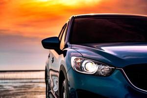 Blue compact SUV car with sport and modern design parked on the beach by the sea at sunset. Environmentally friendly technology. Business success concept. Front view car with open headlamp light. photo