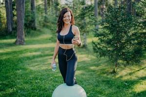 Slim pleased brunette sporty woman leans at fit ball, holds mobile phone, listens music in earphones, downloads songs in playlist, poses with bottle of water, makes exercises in open air at park photo