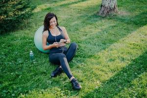 Relaxed sportswoman has rest after doing sport exercises with fitball, uses smartphone, listens favorite music in earphones from playlist, sits on green grass, bottle of fresh water near. Aerobics