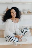 Vertical shot of pleased young Afro American woman feels relaxed and carefree, drinks aromatic coffee, sits crossed legs against kitchen interior, being in good mood, spends spare time at home photo