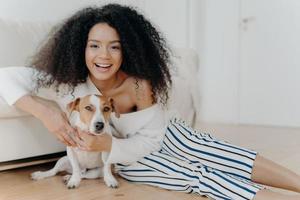 imagen de una mujer feliz y rizada juega con un perrito gracioso, posa en el suelo en una habitación espaciosa, un sofá cerca, sonríe ampliamente, abraza a una mascota con amor, se viste con ropa de moda, se divierte con un animal foto