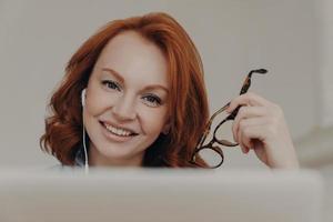 Close up image of ginger positive European woman smiles gently, enjoys online communication, watches tutorial webinar, gets new skills, sits in front of laptop computer, holds transparent glasses photo