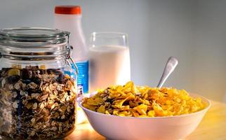 tazón de cereal con cuchara puesto sobre una mesa de madera cerca de granola en un recipiente de vidrio y un vaso de leche. desayuno de calcio para niños antes de ir a la escuela por la mañana. copos de maíz y concepto de leche foto