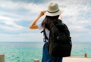 mujer asiática joven feliz en estilo casual con sombrero de paja y mochila. relájese y disfrute de unas vacaciones en la playa del paraíso tropical. chica de pie en el muelle de madera del resort en vacaciones de verano. vibras de verano. foto