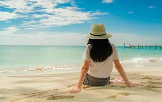 Happy young woman in white shirts and shorts sitting at sand beach. Relaxing and enjoying holiday at tropical paradise beach with blue sky and clouds. Girl in summer vacation. Summer vibes. Happy day. photo