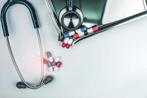 Green stethoscope with pile of antibiotic capsule pills on white table near drug tray. Antimicrobial drug resistance and overuse. Medical equipment for doctor. Clinical Cardiology concept. Health care photo