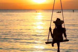 Silhouette woman wear bikini and straw hat swing the swings at the beach on summer vacation at sunset. Girl in swimwear sit on swings and watch beautiful sunset. Summer vibes. Woman travel alone.