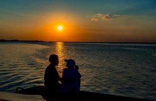 silueta de pareja feliz enamorada sentada y besándose en la playa con cielo naranja y azul al atardecer. vacaciones de verano y concepto de viaje. pareja joven romántica saliendo en la playa. foto