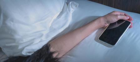mujer asiática durmiendo en la cama en casa y sosteniendo la mano el teléfono móvil. mujer usando teléfono inteligente en el dormitorio. mujer joven adicta al teléfono inteligente. enviar mensajes de texto en el teléfono celular con cansancio hasta quedarse dormido. foto