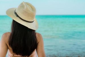 la vista posterior de una joven asiática feliz con traje de baño rosa y sombrero de paja se relaja y disfruta de las vacaciones en la playa del paraíso tropical al atardecer. chica en moda de vacaciones de verano. modelo atractivo de la belleza. vibras de verano. foto