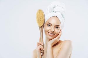Hygiene, beauty, pampering concept. Tender smiling European woman has healthy skin, holds wooden soft massage brush for body, enjoys softness of skin after taking shower, wears wrapped towel. photo