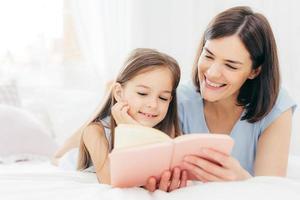 Pretty female mother and her small daughter read interesting book in bedroom, lie on comfortabled bed during morning. Lovely mum reads fairty tale for her little female kid, who is very curious photo