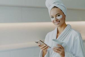 Horizontal shot of glad beautiful womanwith healthy skin cares about undereye skin applies moisturising patches poses in bathrobe uses mobile phone for surfing social networks drinks coffee. photo