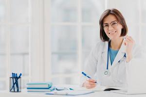 foto de una mujer interna que trabaja en el hospital, escribe en un certificado médico, prescribe un tratamiento para el paciente, hace una receta, se viste con bata blanca con fonendoscopio, deja notas de examen médico