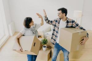 Photo of lovely husband and wife express their agreement, carry cardboard boxes with personal belongings, move in new apartment, pose in empty messy room, decide about interior of living room