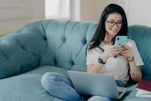 Busy female freelancer does remote job, searches information for presentation, uses modern technologies, surfs internet, sits on comfortable sofa, dressed in casual clothes, creats new publication photo