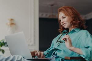 Side view of pretty redhead woman concentrated on remote job, prepares publication for web page, looks gladfully at laptop computer, sits at modern apartment, works remotely, holds eyeglasses photo