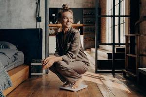 Image of joyful young sitisfied model in satin cozy pajama squatted over scales with wide smile in modern bedroom photo