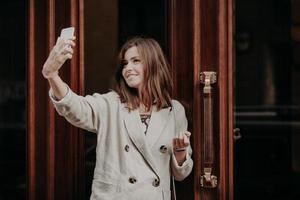 Adorable female student poses for making selfie, uses modern cell phone, dressed in white jacket, stands near entrance to building, has happy expression. People, modern technologies concept. photo