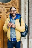 Bearded traveler with charming smile wearing trendy clothes and holding backpack standing indoors in gallery looking into distance messaging with relatives, reading sms. Stylish man having holidays photo