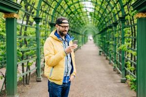 Trendy guy with beard and moustache wearing anorak, jeans and cap standing sideways having attentive look into his smartphone isolated over green arch. Human, style and modern technology concept photo