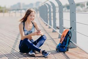 Outdoor shot of delighted pretty female sits crossed legs, uses modern cell phone for listening music and messaging with friends, takes break after jogging exercise, uses free internet connection