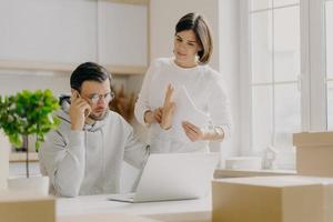 Serious male calls via smartphone, makes refusal gesture, wears spectacles and sweatshirt, asks wife to wait, tries to decide financial problems, pose in kitchen with big windows surrounded with boxes photo