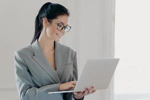 Self confident cheerful woman economist analyzes report about company income on laptop computer, shares multimedia files, has dark hair combed in pony tail, dressed in formal outfit, stands indoor photo