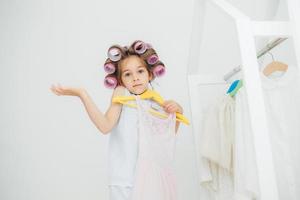 Hesitant girl with clueless expression, doesnt know what to wear, holds hangers with dress, has curlers on head, isolated over white background. Cute child with pleasant appearance prepares for party photo