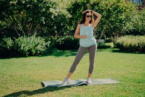 Fitness sport and recreation concept. Full length shot of sporty woman dressed in t shirt leggings and sneakers stands on karemat holds bottle of water poses against green nature background. photo