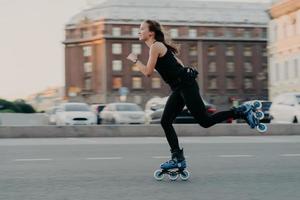 mujer atlética monta en rodillos se mueve muy rápido vestida con ropa activa disfruta de patinar siendo fotografiada en poses de acción en un lugar urbano dedicado a deportes extremos. concepto de estilo de vida activo foto