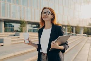 Dreamy redhead woman in formal clothes carries notepad tablet disposable cup of coffee photo