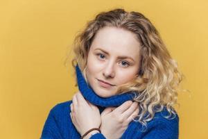 Candid shot of serious adorable woman with curly hair, wears warm blue sweater, warms herself after walk outdoor, isolated over yellow studio background. Attractive female has confident expression photo