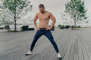 Outside of athlete guy does sport exercises in fresh air, stands with naked torso, has muscular body and stands outside. Active unshaven guy in good physical shape, has regular workout in morning photo