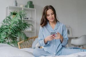 Photo of relaxed young European lady in pyjamas enjoys audio playlist, listens music romantic songs in earphones, uses modern cell phone, sits on comfortable bed against domestic atmosphere.