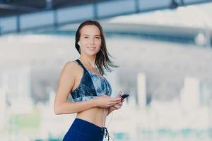 Active Caucasian woman with dark hair, healthy skin, dressed in sportswear, uses up to date gadget, listens music with earphones, has charming smile, stands outdoor. People and lifestyle concept photo