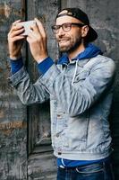 Excited male with beard and moustache wearing trendy denim shirt with hook and jeans looking happily into camera of his smartphone making selfie. Wonderful hipster man using modern device isolated photo