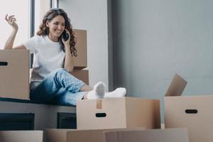 Happy hispanic woman sitting on windowsill of new house. Girl unpacking boxes and talking on phone. photo