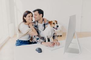 Affectionate couple in love embrace and going to kiss, watch romantic movie at modern computer, pedigree dog focused in monitor with interest. Lovely family spend free time in domestic atmosphere photo