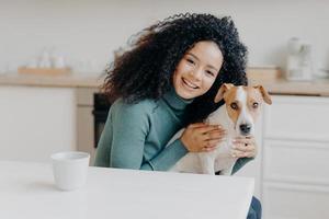 Adorable smiling woman embraces her favourite dog, expresses care and love, good attitiude to pet, poses against kitchen interior, drinks aromatic beverage. People, animals, relationship, affection photo