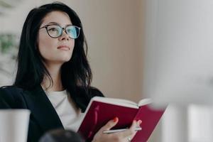 Beautiful woman with thoughtful expression, holds opened notepad, writes time management, makes plans for productive day, wears eyeglasses, poses indoor. Self employed female writes down ideas photo