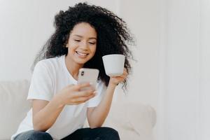 Peaceful curly woman scrolling social networks via modern mobile phone, watches funny videos, views photos in social networks, drinks coffee, poses on couch in living room, gets high paid job offer