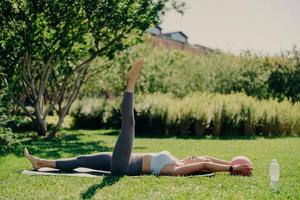 Outdoor shot of active fit woman does aerobics exercises with fitness ball lying on karemat raises leg poses on green grass with closed eyes being alone with nature enjoys tranquil atmosphere photo