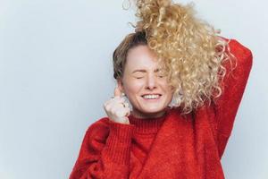 Lucky young cheerful female feels excited, keeps eyes shut and clenches fists, has curly light pony tail, being overjoyed as recieves victory in competition, isolated over white studio background photo