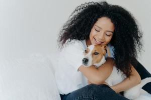 Happy African American woman expresses love to dog, embraces pet, dressed in casual wear, sit on comfortable bed in bedroom, sleeps with domestic animal. Jack russell terrier with owner in morning photo