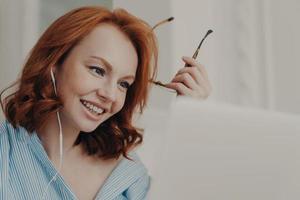 Close up shot of redhead businesswoman makes video call to business partner, has discussion with corporate partner, holds optical glasses, smiles pleasantly, uses earphones. Remote job interview
