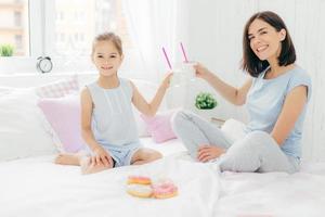 Horizontal shot of pretty female and her daughter clink glasses with cocktail in bed, have breakfast in bedroom, eat delicious doughnuts, look happily at camera. Young mother and her kid at home photo